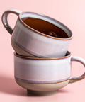 Two stacked ceramic mugs with a dark liquid on a pink background