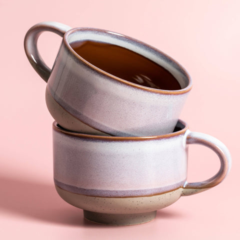 Two stacked ceramic mugs with a dark liquid on a pink background