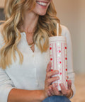 A woman in a white ribbed cardigan holds a white insulated tumbler with a red and white fall pattern and a straw in a softly lit indoor setting.