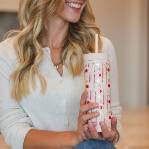 A woman in a white ribbed cardigan holds a white insulated tumbler with a red and white fall pattern and a straw in a softly lit indoor setting.