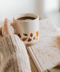 Person holding a mug with a leaf pattern next to a book on a light surface.