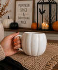 A hand with orange and brown nail polish holds a pumpkin shaped ceramic mug with a handle, set on a burlap placemat in a cozy fall-themed room with pumpkins and a framed sign.