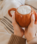 Person holding a pumpkin mug with a hot beverage and whipped cream, wearing a beige sweater.