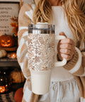 Person holding a mug with a leaf pattern in a cozy indoor setting with pumpkins and candles.