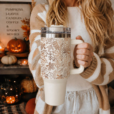 Person holding a mug with a leaf pattern in a cozy indoor setting with pumpkins and candles.