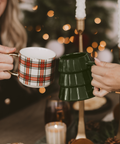 Two people holding mugs in front of a decorated Christmas tree with lights.
