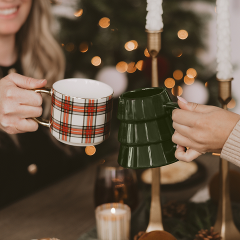 Two people holding mugs in front of a decorated Christmas tree with lights.