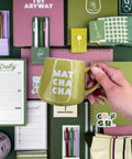 A hand holds a green ceramic mug with the words 'MATH CHA CHA' printed in white. The mug is surrounded by coordinated stationery items including notebooks, pens, sticky notes, and a sock on a flat-lay background.