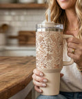 A woman in a kitchen holds a large insulated tumbler with a handle, clear lid, and straw. The tumbler has a warm cream body with a copper-toned leaf pattern on the upper section.