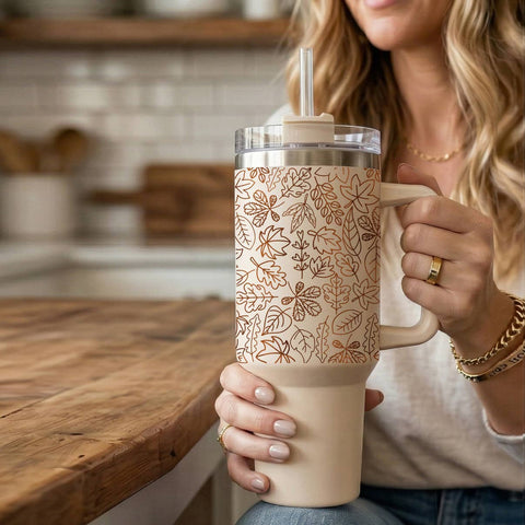 A woman in a kitchen holds a large insulated tumbler with a handle, clear lid, and straw. The tumbler has a warm cream body with a copper-toned leaf pattern on the upper section.
